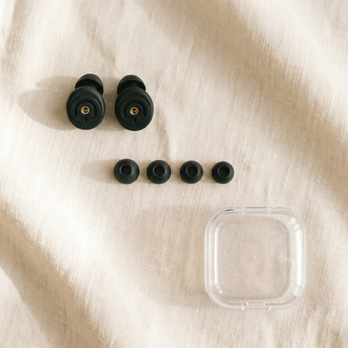 A pair of round black silicone earplugs, four ear tips and a clear case on linen
