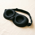 A black contoured sleep mask with molded eye cups on a pale sheet