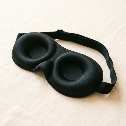 A black contoured sleep mask with molded eye cups on a pale sheet