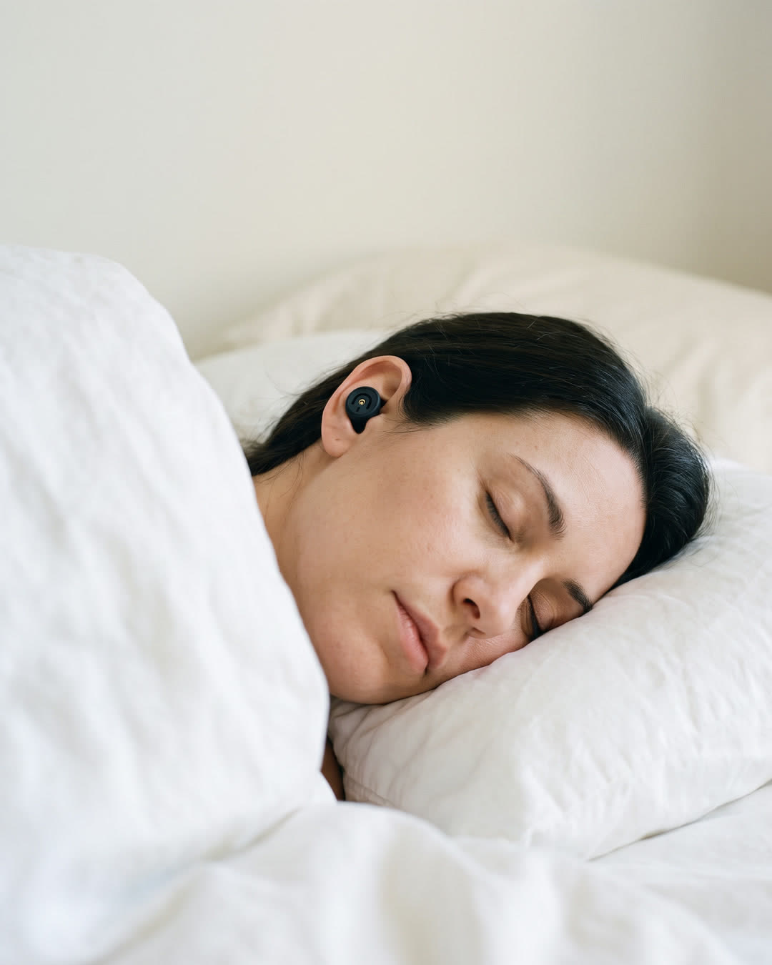 A woman asleep on her side under a white duvet, a small black earplug in her ear